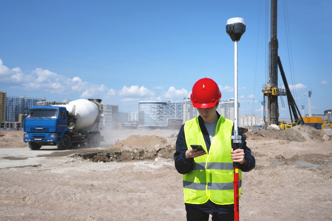 Surveyor in hard hat and safety vest checking phone next to GNSS survey pole on a construction site